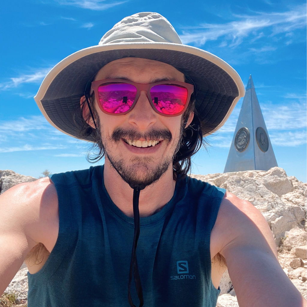 Justin Langhorst in sunglasses and a hat at Guadalupe Peak in Guadalupe Mountains National Park with monument in the background
